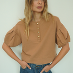 A woman modeling a cropped tan top with puff sleeves and gold-tone buttons down the front, paired with blue jeans, against a plain light background.