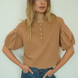 A woman modeling a cropped tan top with puff sleeves and gold-tone buttons down the front, paired with blue jeans, against a plain light background.