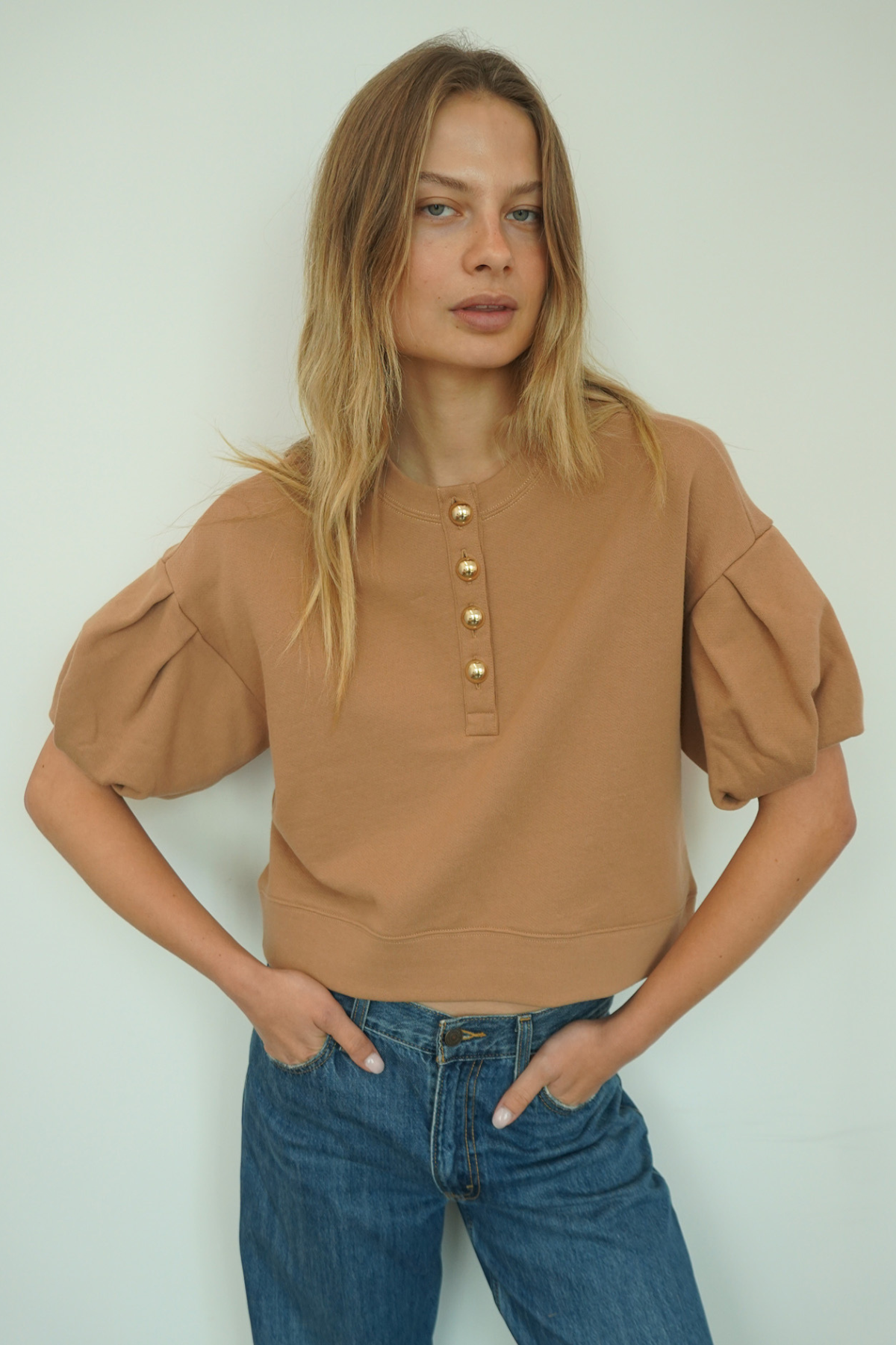 A woman modeling a cropped tan top with puff sleeves and gold-tone buttons down the front, paired with blue jeans, against a plain light background.