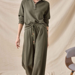 A woman modeling a matching olive green ribbed knit lounge set consisting of a long-sleeve collared top and wide-leg pants, styled with a drawstring waist. She is standing against a draped beige fabric backdrop.