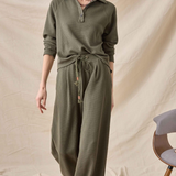 A woman modeling a matching olive green ribbed knit lounge set consisting of a long-sleeve collared top and wide-leg pants, styled with a drawstring waist. She is standing against a draped beige fabric backdrop.