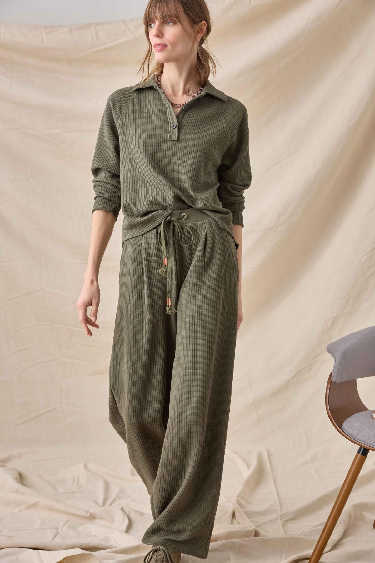A woman modeling a matching olive green ribbed knit lounge set consisting of a long-sleeve collared top and wide-leg pants, styled with a drawstring waist. She is standing against a draped beige fabric backdrop.