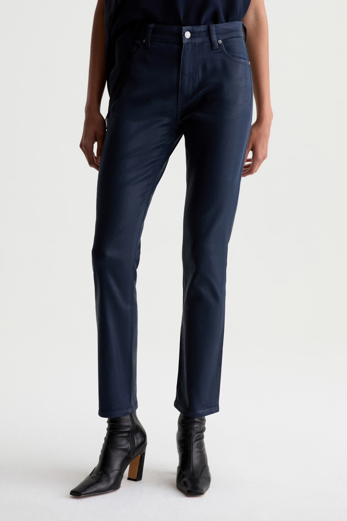 A person wearing dark navy slim-fit jeans with black pointed-toe ankle boots, shown from the waist down against a plain light gray studio background.