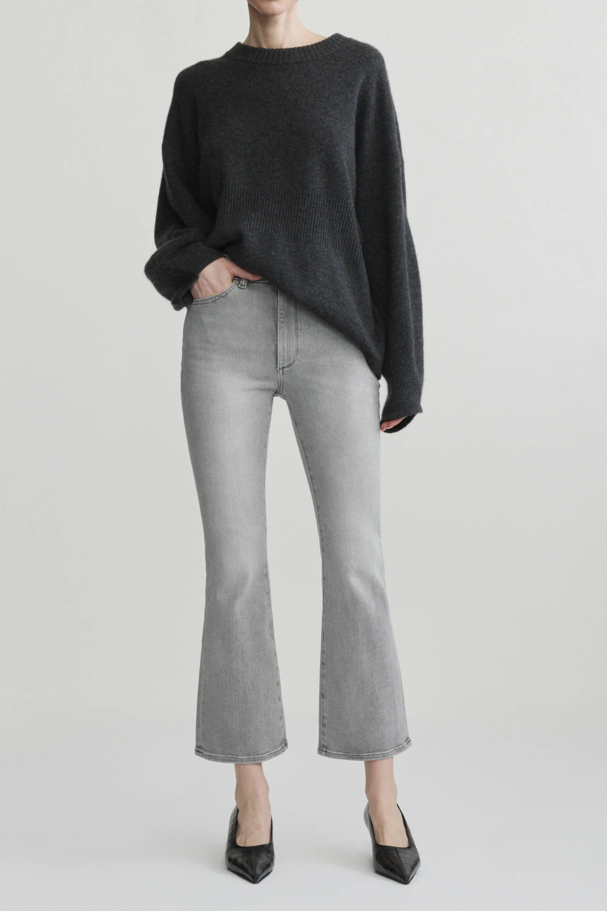 A model wearing a dark charcoal gray knit sweater, light gray cropped flare jeans, and black pointed-toe heels, standing against a light gray studio background.