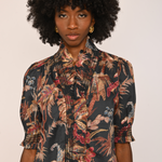 A model with a large black afro wearing a short-sleeved button-up blouse with a dark tropical floral print in red, beige, and brown, paired with dark brown velvet pants, against a plain light beige background.