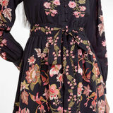 A close-up of a woman wearing a dark navy floral dress with pink, red, and green botanical patterns, featuring a V-neckline, button front, and self-tie waist belt.