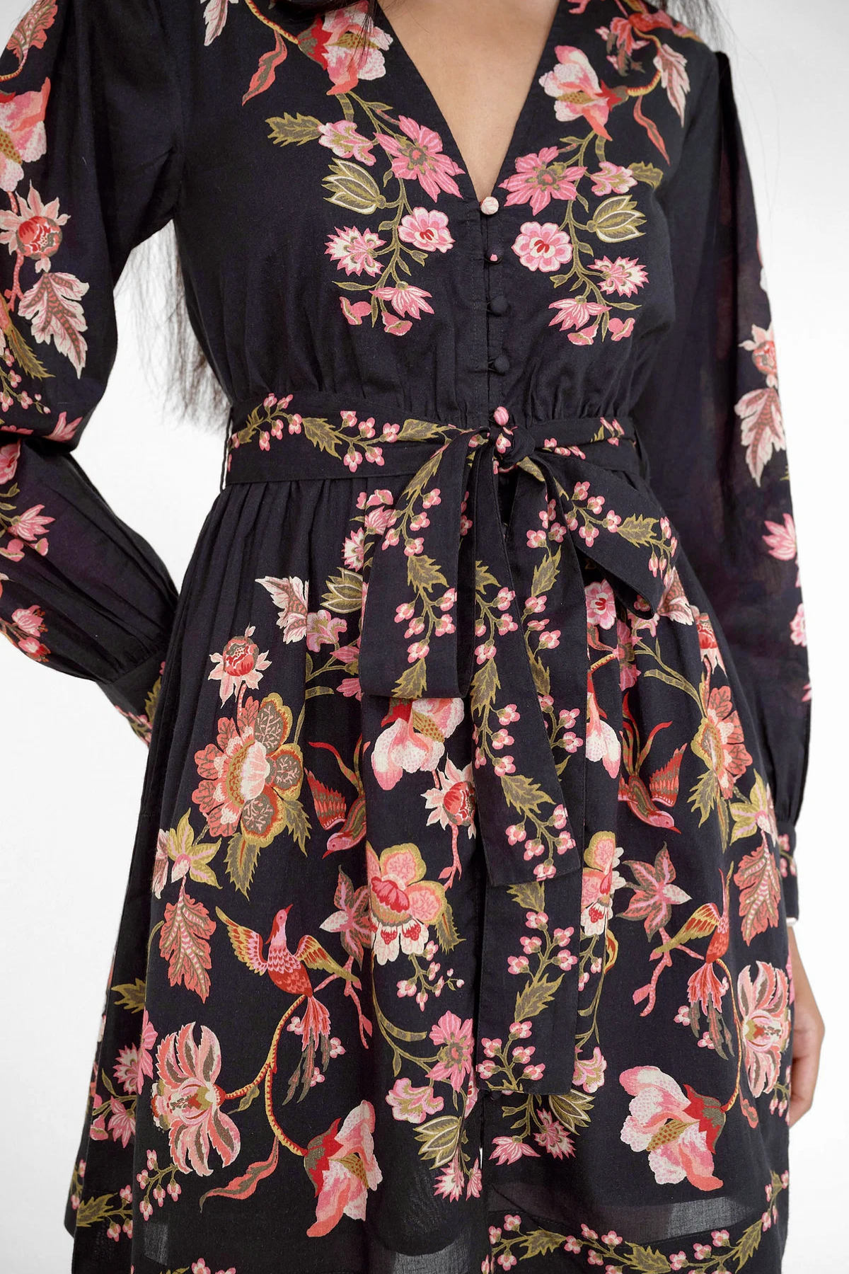 A close-up of a woman wearing a dark navy floral dress with pink, red, and green botanical patterns, featuring a V-neckline, button front, and self-tie waist belt.