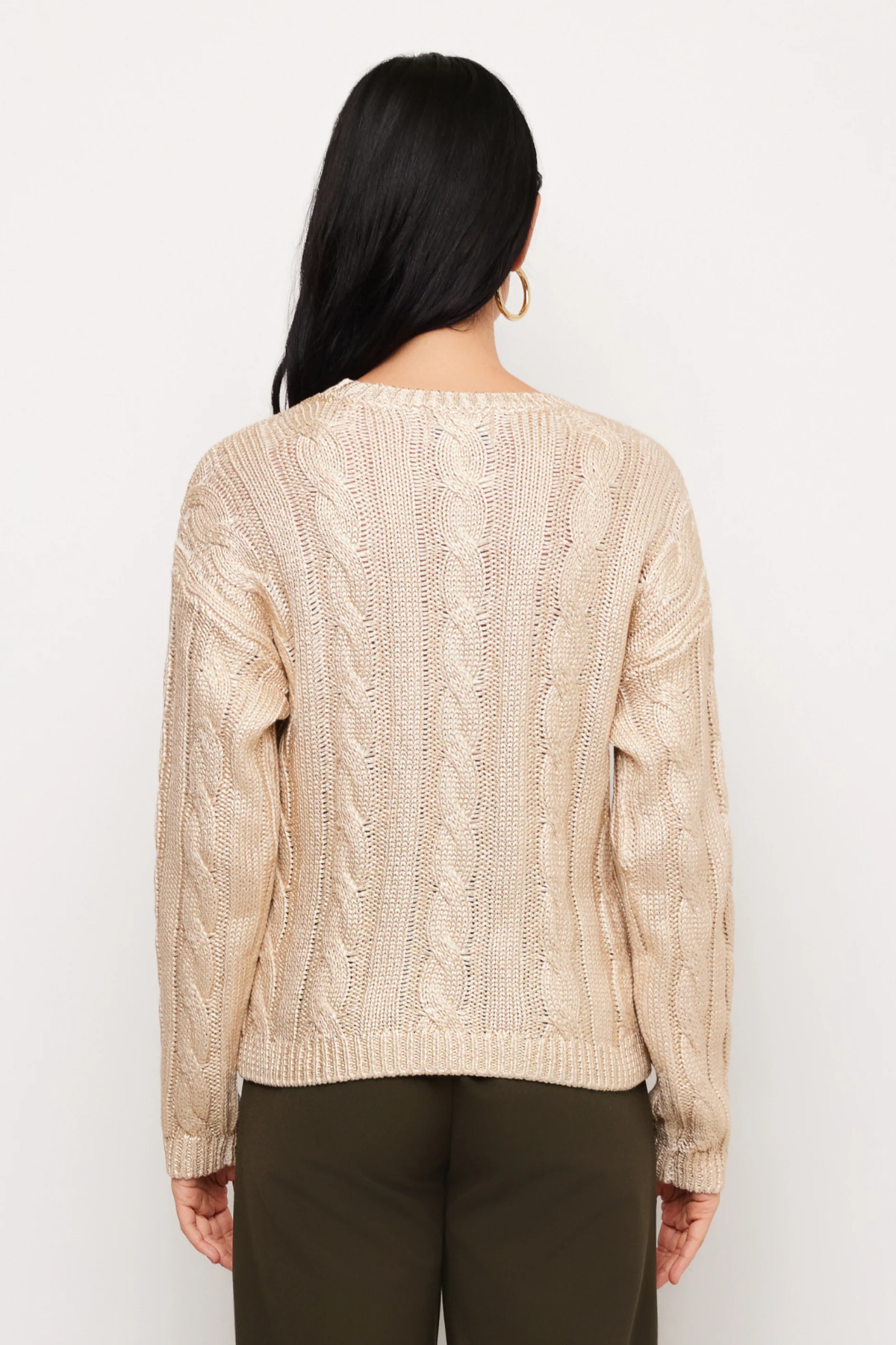 A woman viewed from the back wearing a cream cable-knit sweater and dark olive pants against a plain white background.