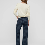 A woman is shown from behind wearing a cream-colored long-sleeve top and dark blue patterned flared pants against a light gray studio background.