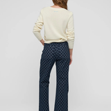 A woman is shown from behind wearing a cream-colored long-sleeve top and dark blue patterned flared pants against a light gray studio background.