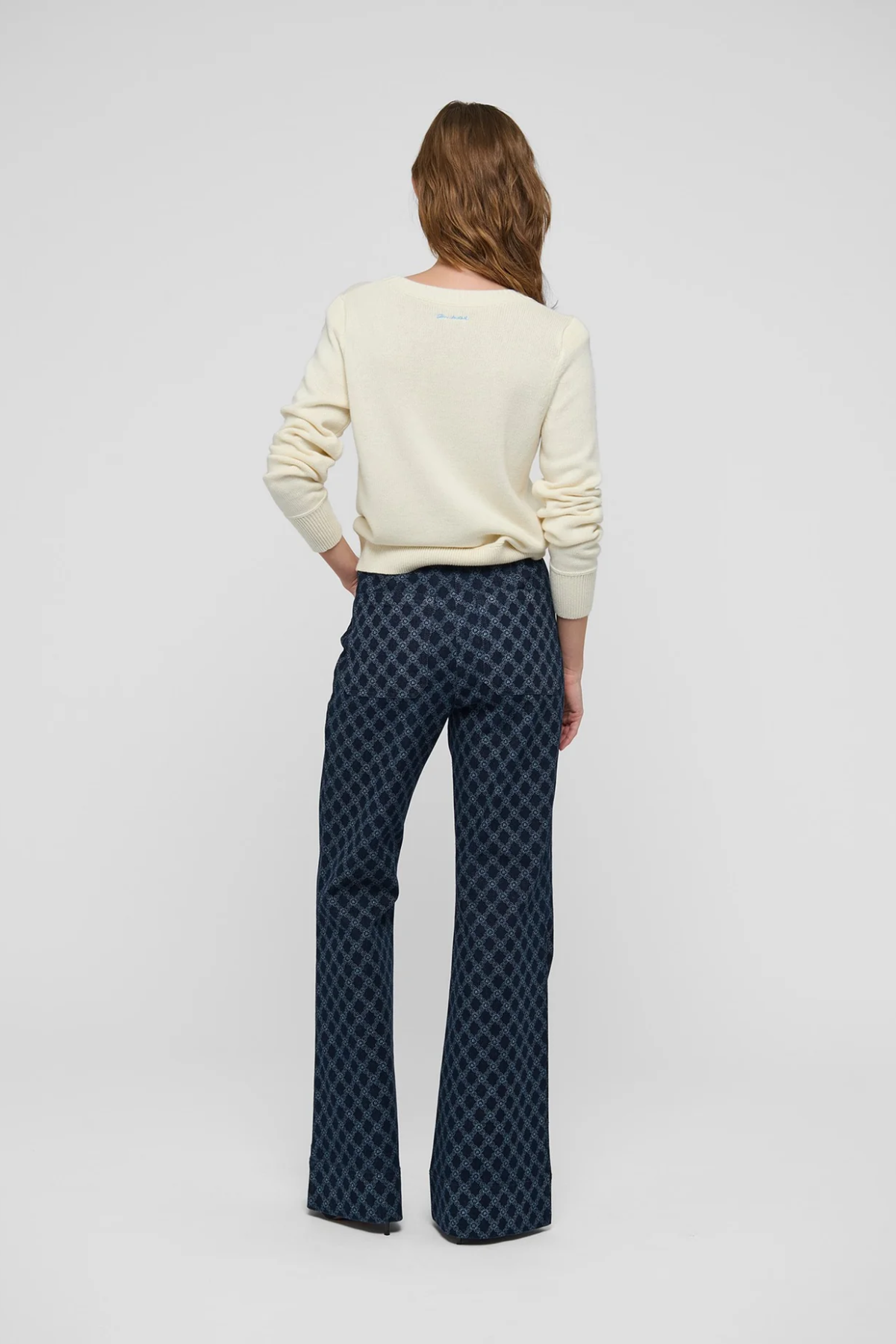 A woman is shown from behind wearing a cream-colored long-sleeve top and dark blue patterned flared pants against a light gray studio background.