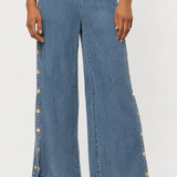 A model wearing high-waisted, wide-leg blue denim pants with decorative gold-tone buttons running down the outer sides, paired with metallic heeled sandals against a plain white studio background.