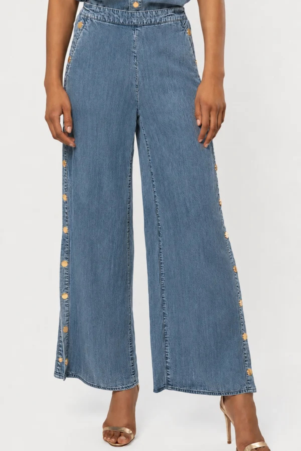 A model wearing high-waisted, wide-leg blue denim pants with decorative gold-tone buttons running down the outer sides, paired with metallic heeled sandals against a plain white studio background.