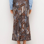 A person shown from the back wearing a brown batik-patterned midi skirt with blue, beige, and white details, paired with a light blue shirt and black block-heel mules, against a plain white studio background.