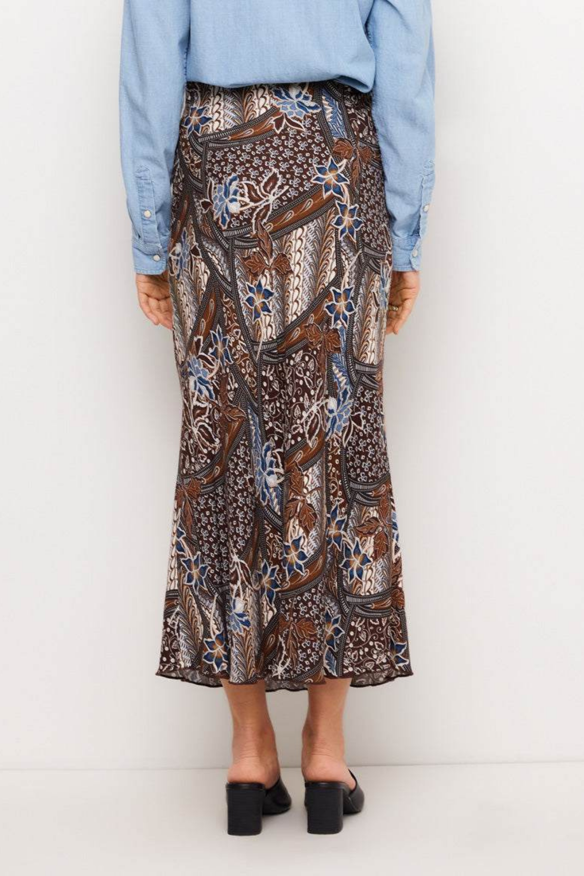 A person shown from the back wearing a brown batik-patterned midi skirt with blue, beige, and white details, paired with a light blue shirt and black block-heel mules, against a plain white studio background.