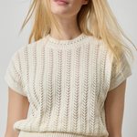 A blonde woman wearing a cream-colored short-sleeve knit sweater with a textured vertical cable pattern, paired with dark olive green pants, against a light gray studio background.