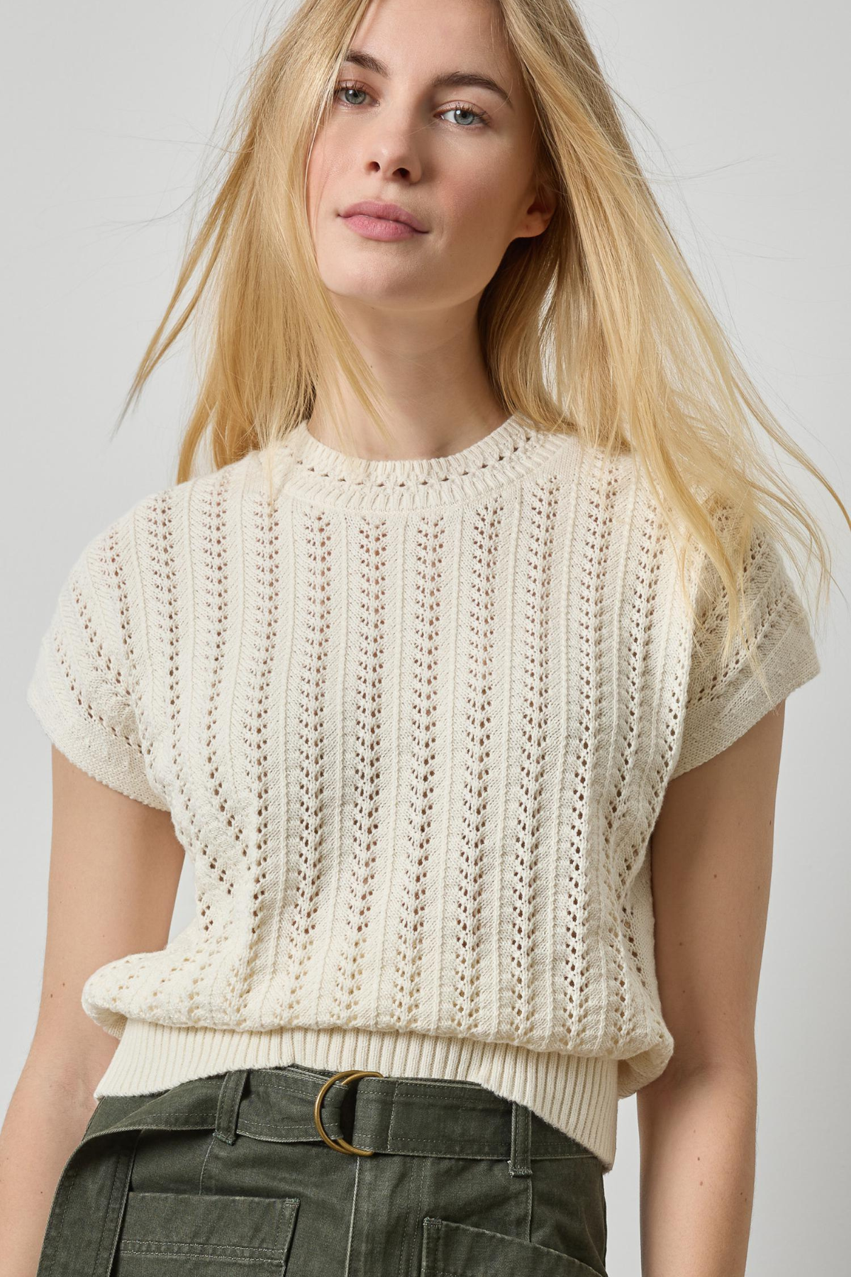 A blonde woman wearing a cream-colored short-sleeve knit sweater with a textured vertical cable pattern, paired with dark olive green pants, against a light gray studio background.