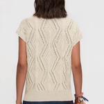 A woman viewed from the back wearing a cream-colored short-sleeve knit top with a textured geometric diamond pattern and dark blue pants, set against a plain light gray background.