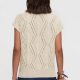 A woman viewed from the back wearing a cream-colored short-sleeve knit top with a textured geometric diamond pattern and dark blue pants, set against a plain light gray background.