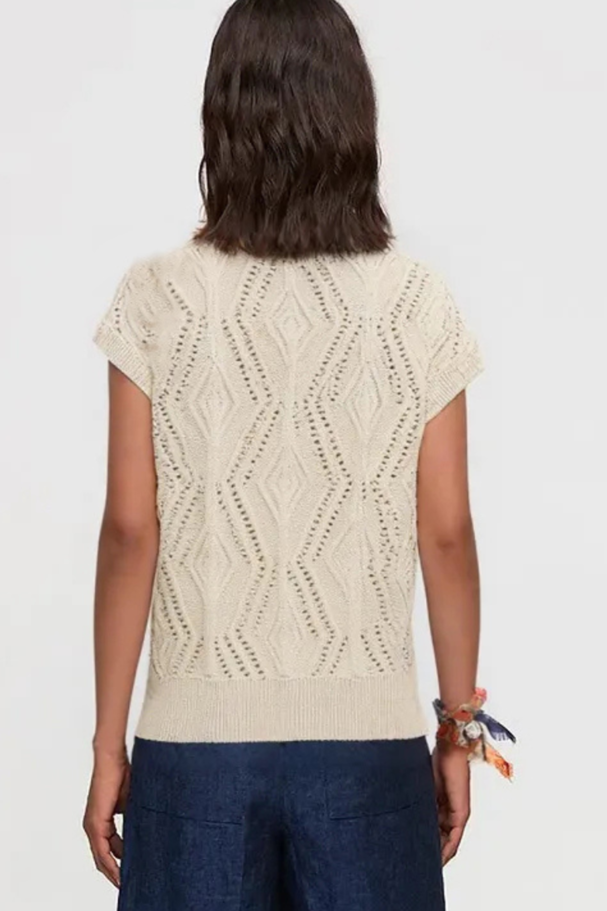 A woman viewed from the back wearing a cream-colored short-sleeve knit top with a textured geometric diamond pattern and dark blue pants, set against a plain light gray background.