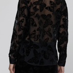 A model shown from the back wearing a sheer black long-sleeve blouse with a black embroidered butterfly pattern, paired with black trousers, against a light gray studio background.