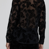 A model shown from the back wearing a sheer black long-sleeve blouse with a black embroidered butterfly pattern, paired with black trousers, against a light gray studio background.