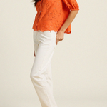 side of woman in orange eyelet top and white denim on tan background 