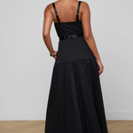 A woman viewed from the back wearing a black sleeveless maxi dress with thin shoulder straps, a structured upper bodice, a thin waistband detail, and a full gathered skirt. She is standing on a light wood floor against a plain light gray wall.