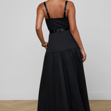 A woman viewed from the back wearing a black sleeveless maxi dress with thin shoulder straps, a structured upper bodice, a thin waistband detail, and a full gathered skirt. She is standing on a light wood floor against a plain light gray wall.