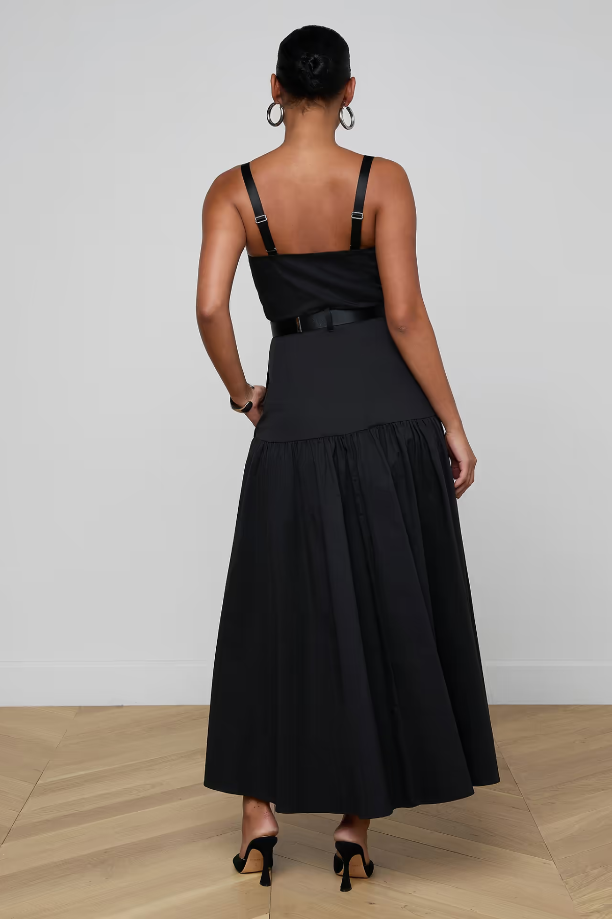 A woman viewed from the back wearing a black sleeveless maxi dress with thin shoulder straps, a structured upper bodice, a thin waistband detail, and a full gathered skirt. She is standing on a light wood floor against a plain light gray wall.