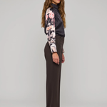 A woman stands in profile wearing a dark charcoal turtleneck top with long floral sleeves, dark brown wide-leg trousers, and dark pointed flats against a light gray studio background.