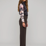 A woman stands in profile wearing a dark charcoal turtleneck top with long floral sleeves, dark brown wide-leg trousers, and dark pointed flats against a light gray studio background.