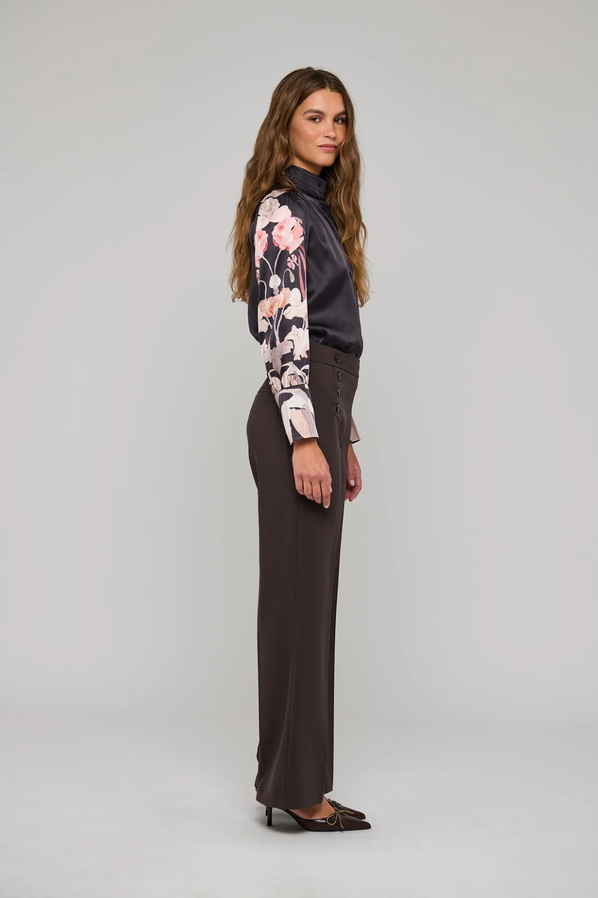 A woman stands in profile wearing a dark charcoal turtleneck top with long floral sleeves, dark brown wide-leg trousers, and dark pointed flats against a light gray studio background.