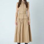 A woman modeling a sleeveless beige midi dress with a structured collar, front button placket, matching belt, and flared skirt, paired with black pointed boots against a light gray studio background.