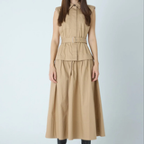 A woman modeling a sleeveless beige midi dress with a structured collar, front button placket, matching belt, and flared skirt, paired with black pointed boots against a light gray studio background.