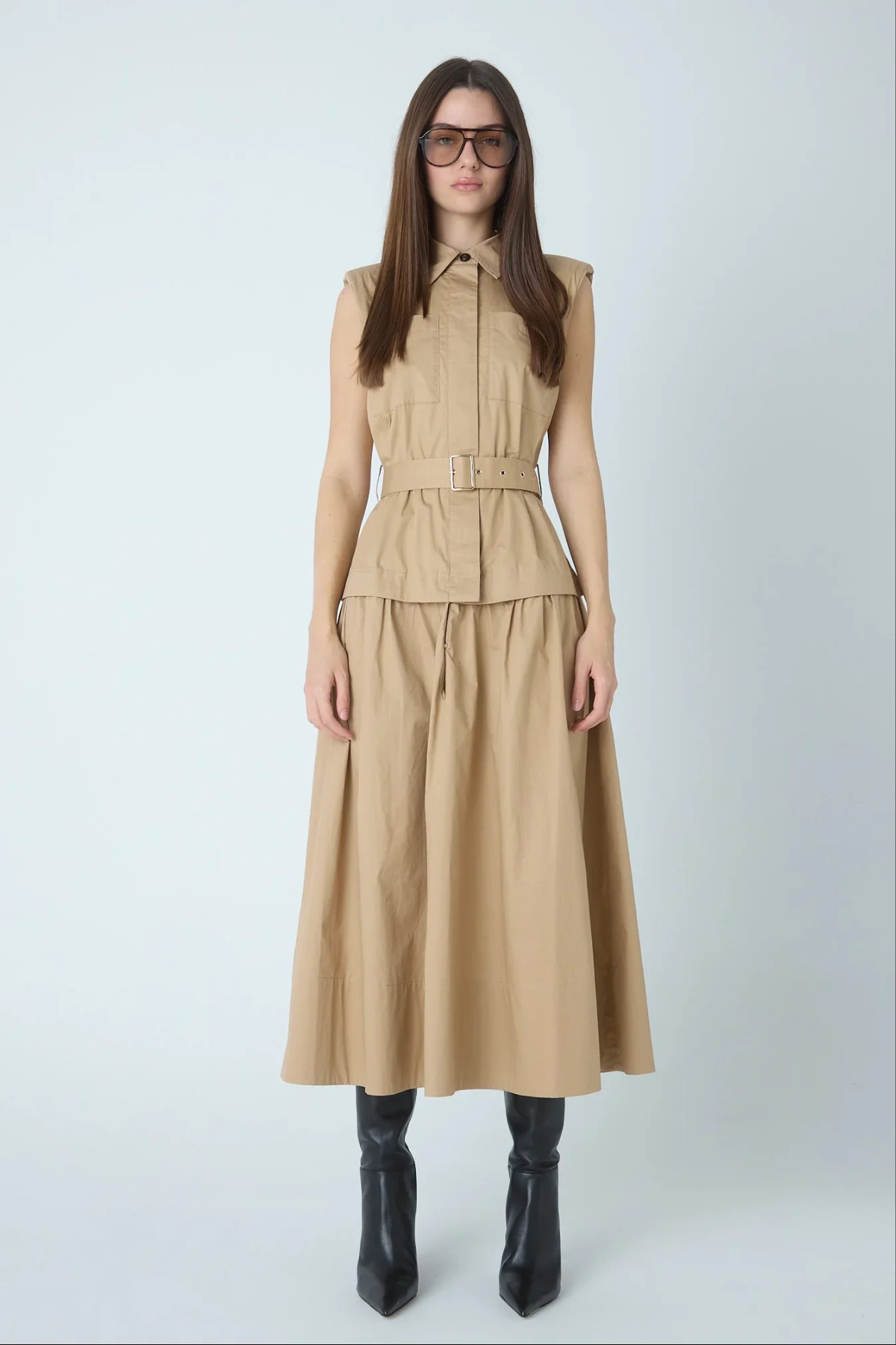 A woman modeling a sleeveless beige midi dress with a structured collar, front button placket, matching belt, and flared skirt, paired with black pointed boots against a light gray studio background.