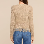 A woman viewed from the back wearing a textured beige cropped sweater with fringe trim and blue jeans, against a plain beige studio background.