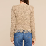 A woman viewed from the back wearing a textured beige cropped sweater with fringe trim and blue jeans, against a plain beige studio background.