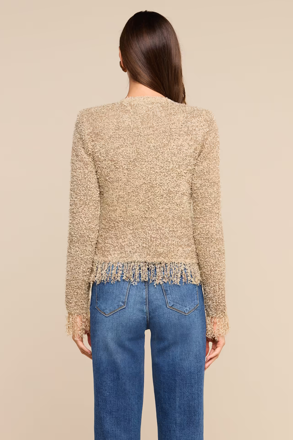A woman viewed from the back wearing a textured beige cropped sweater with fringe trim and blue jeans, against a plain beige studio background.