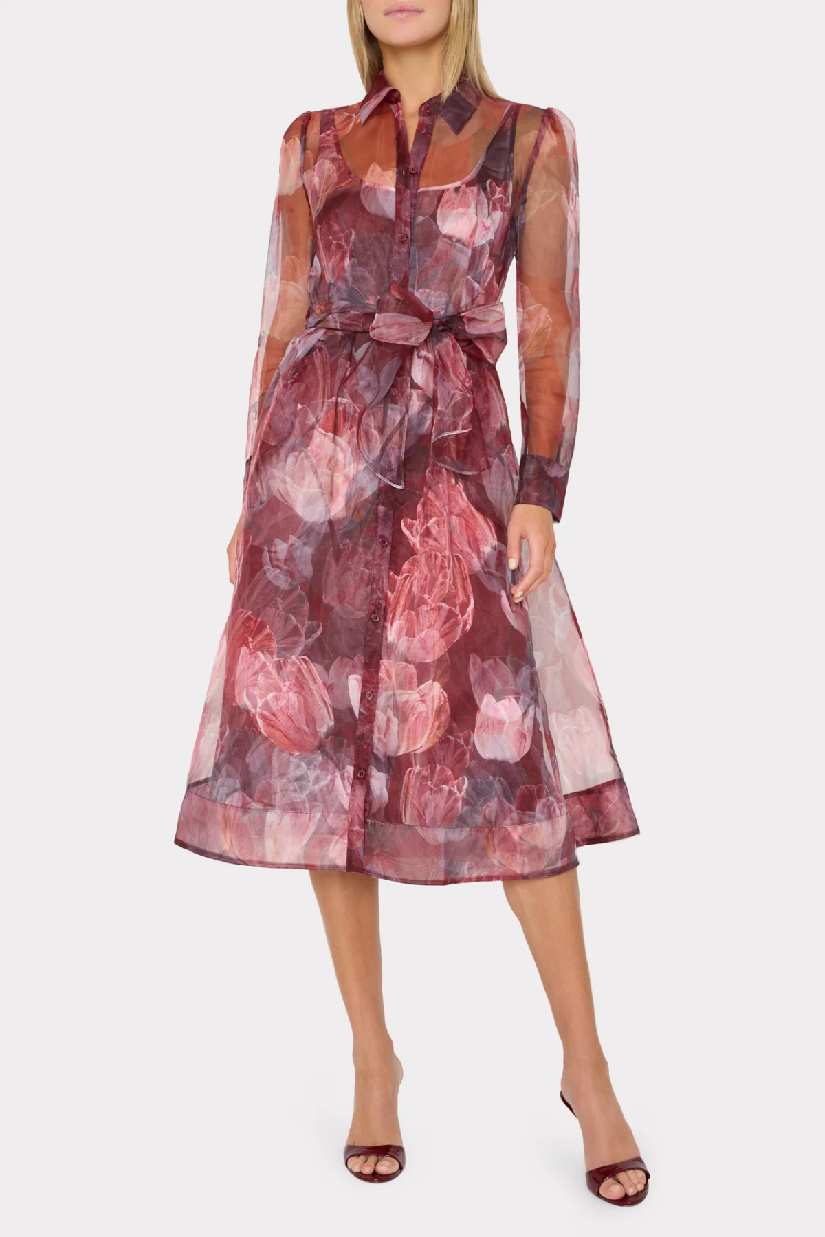 A woman models a sheer long-sleeve midi dress with a floral print in shades of dusty rose, burgundy, and mauve, featuring a tie waist and a collared neckline, set against a plain light gray background.