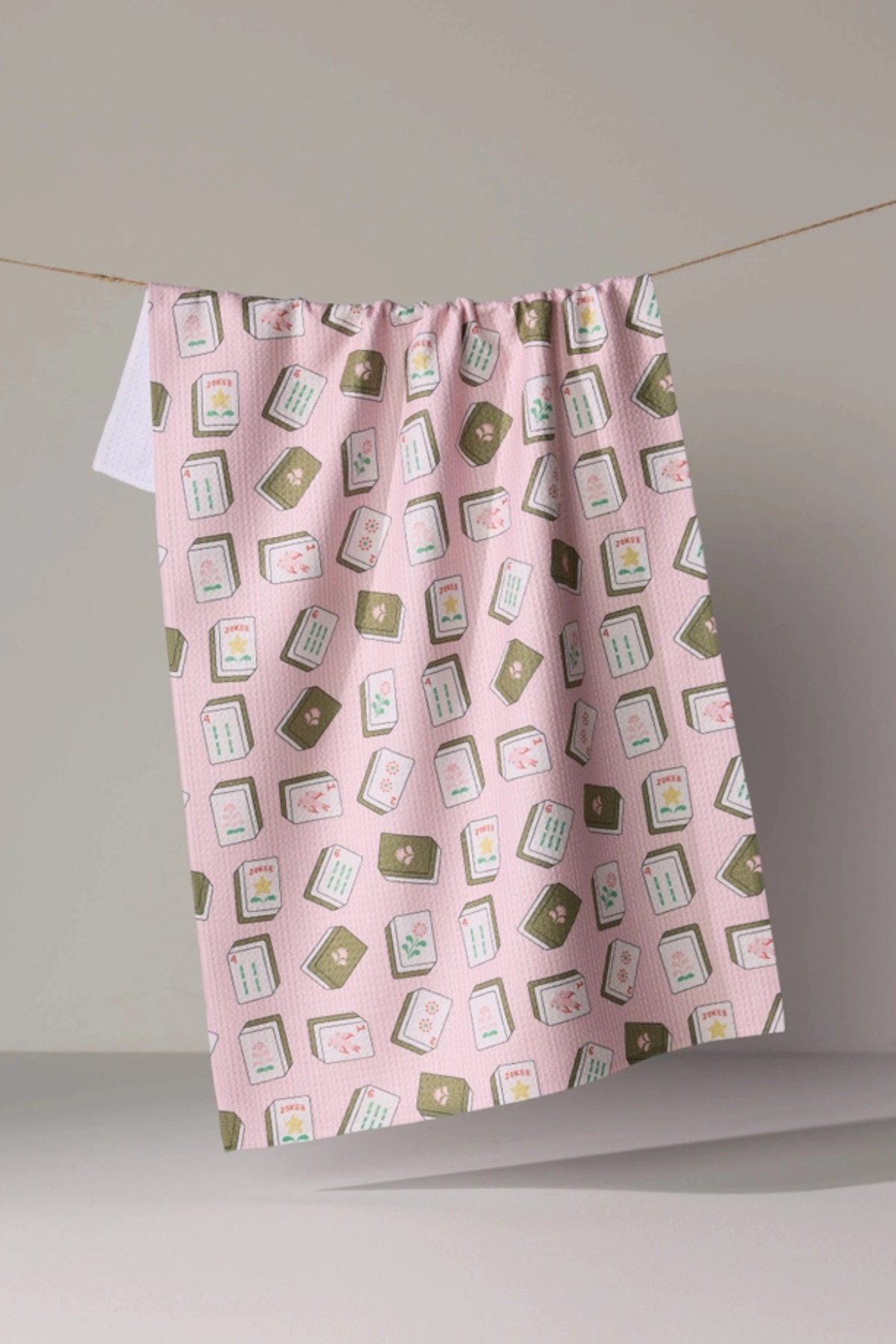 A pink kitchen towel with a repeating pattern of illustrated playing cards, hanging from a string against a neutral gray background.
