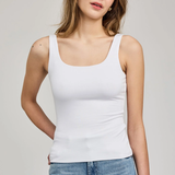A woman wearing a fitted white scoop-neck tank top and light blue jeans, standing against a plain light gray studio background.