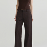 A model wearing a sleeveless dark brown mock-neck top and dark brown pinstriped wide-leg trousers, paired with dark pointed-toe heels, against a light gray studio background.