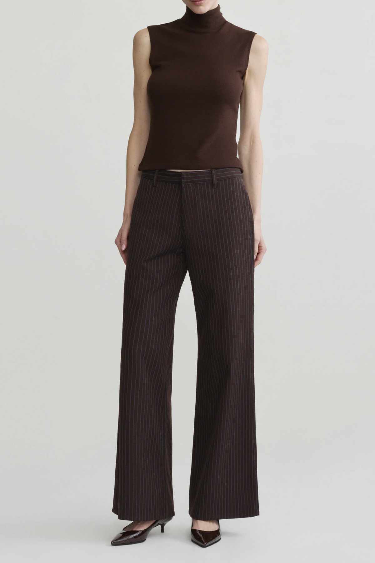 A model wearing a sleeveless dark brown mock-neck top and dark brown pinstriped wide-leg trousers, paired with dark pointed-toe heels, against a light gray studio background.