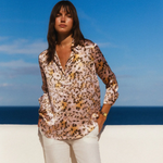 Woman in a patterned blouse and white pants standing on a beach with blue sky and ocean in the background