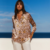 Woman in a patterned blouse and white pants standing on a beach with blue sky and ocean in the background