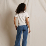 A model viewed from behind wearing blue wide-leg jeans and a white t-shirt, standing against a draped cream fabric backdrop.