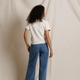 A model viewed from behind wearing blue wide-leg jeans and a white t-shirt, standing against a draped cream fabric backdrop.