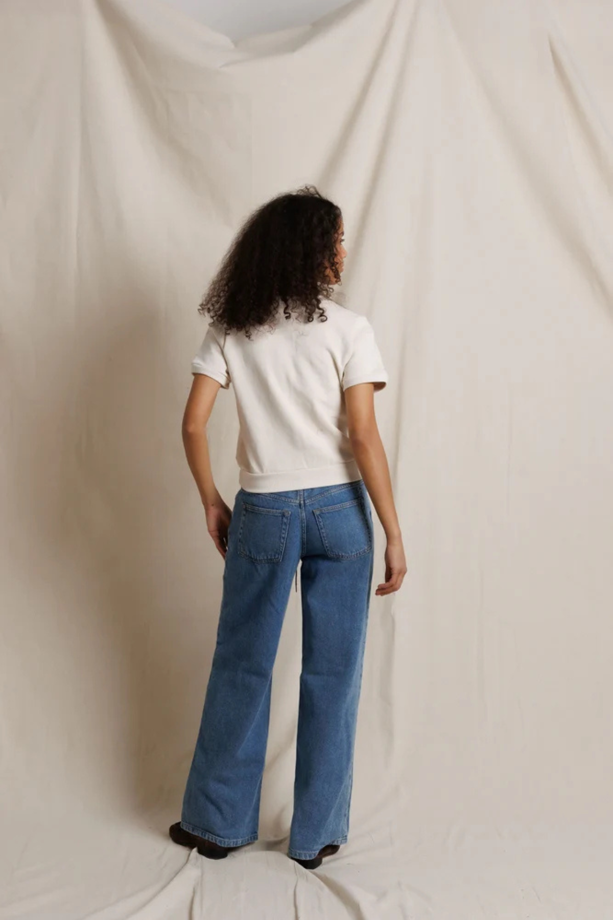 A model viewed from behind wearing blue wide-leg jeans and a white t-shirt, standing against a draped cream fabric backdrop.