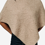A woman wearing a beige knit poncho sweater with a textured geometric diamond pattern, paired with dark blue jeans, shown against a plain light gray background.